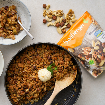 Pfanne mit Apfel-Crumble und Vanilleeis, daneben verpackte Nussmischung und Schüssel auf Tisch.