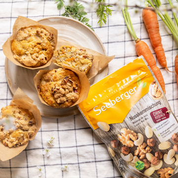 Muffins auf Teller ruhen neben Seeberger Nusspackung, dekoriert mit Karotten auf Tischdecke.