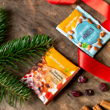 Zwei Seeberger Snack-Tüten liegen neben Tannenzweigen, roten Beeren und Zimtstangen auf Holz.