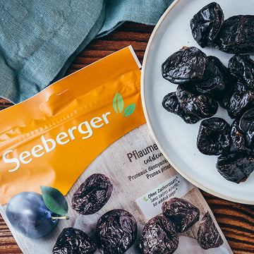 Pflaumen auf Teller, neben gelber Seeberger-Verpackung, auf hölzernem Tisch mit blauer Serviette.